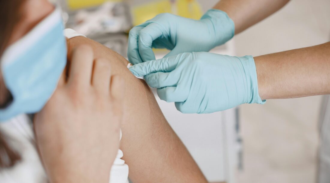 A person wearing medical gloves applies a bandaid on the shoulder of a person wearing a mask
