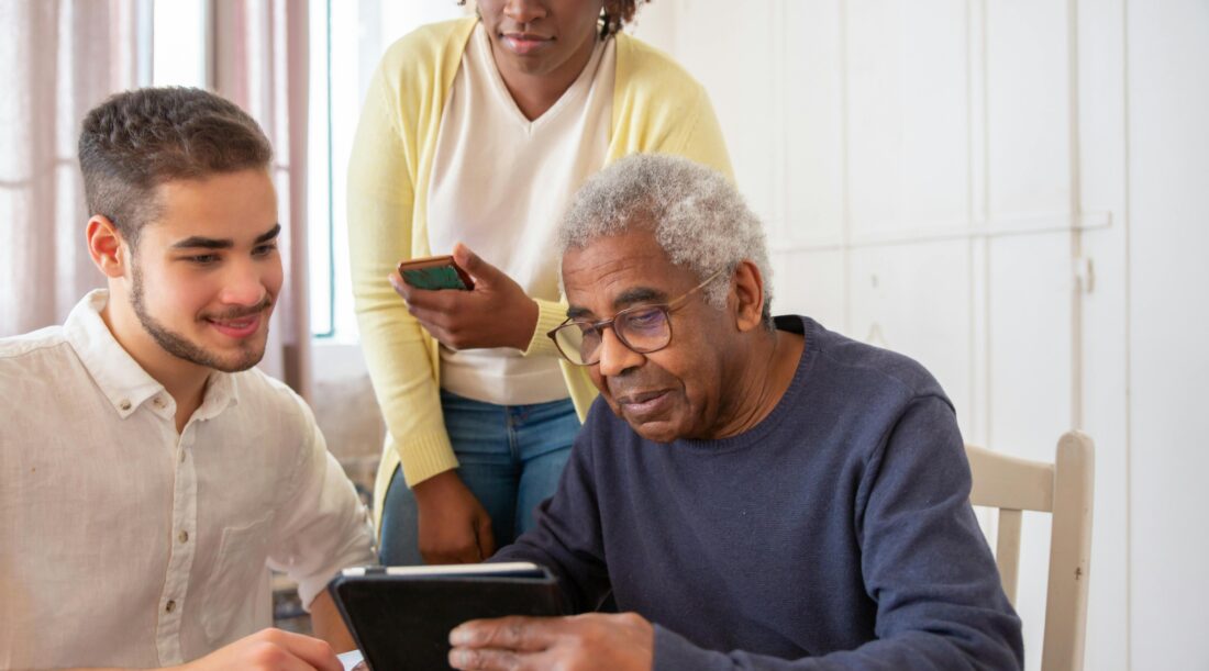 An older man holds a tablet while talking to two younger people