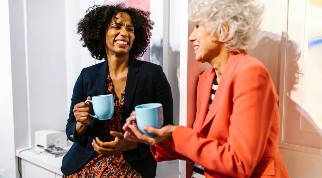 Two older women hold coffee cups while smiling at each other