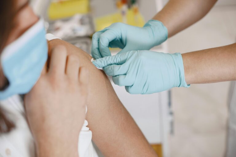 A person wearing medical gloves applies a bandaid on the shoulder of a person wearing a mask