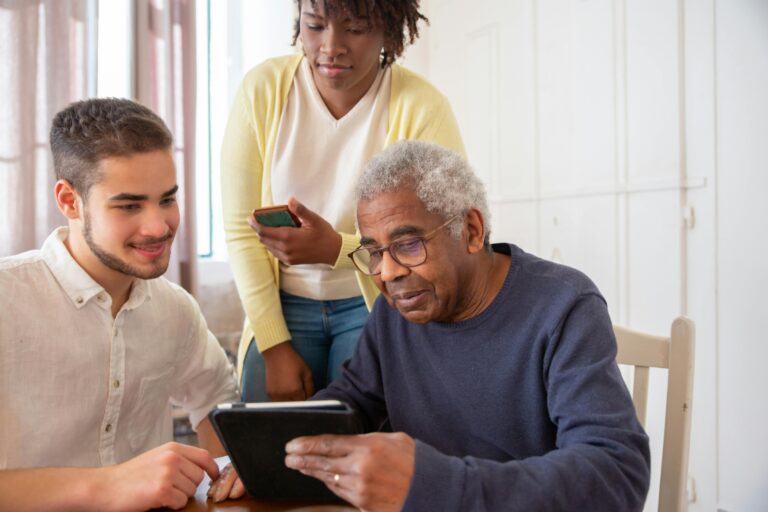 An older man holds a tablet while talking to two younger people