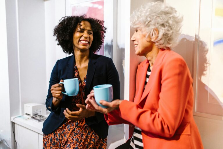 Two older women hold coffee cups while smiling at each other